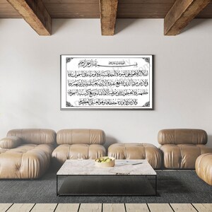 May include: A framed black and white print of Arabic calligraphy with a decorative border. The print is hanging on a white wall in a living room with a brown leather sofa and a coffee table.