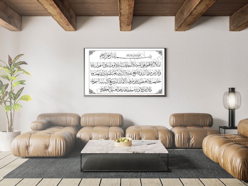 May include: A framed black and white print of Arabic calligraphy with a decorative border. The print is hanging on a white wall in a living room with a brown leather sofa and a coffee table.