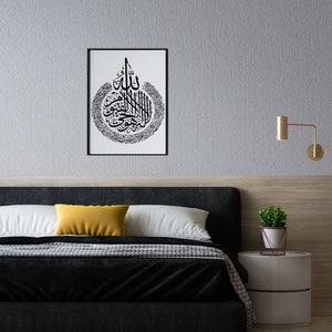May include: A black and white framed print of Islamic calligraphy in a bedroom setting. The print is hanging on a gray wall above a black headboard with white and yellow pillows. A small potted plant sits on a white bedside table.