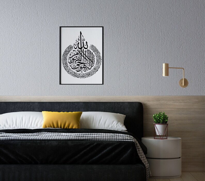 May include: A black and white framed print of Islamic calligraphy in a bedroom setting. The print is hanging on a gray wall above a black headboard with white and yellow pillows. A small potted plant sits on a white bedside table.
