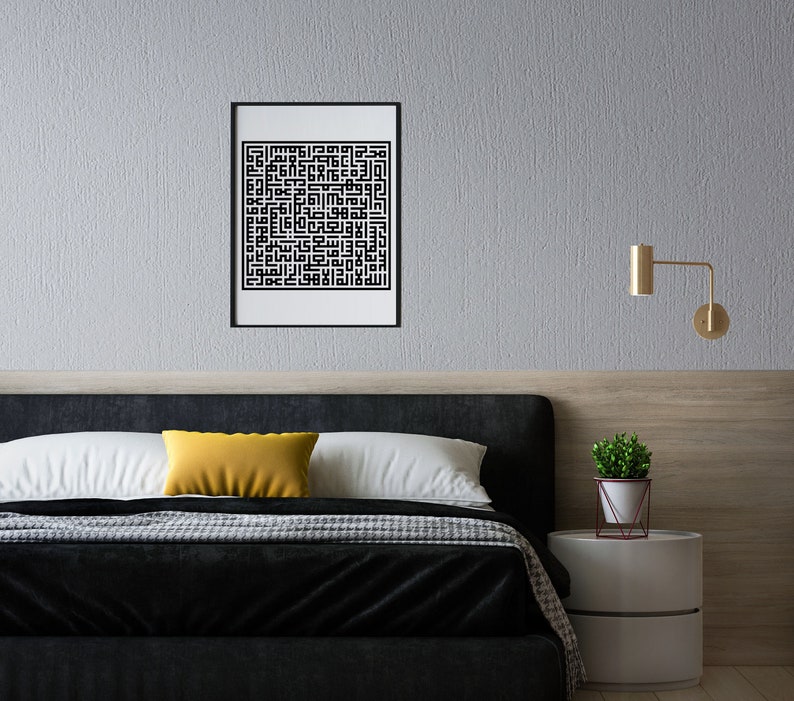 May include: A black and white framed print of Islamic calligraphy hangs on a gray wall above a black headboard bed with a yellow pillow. A small potted plant sits on a white nightstand with a gold wall sconce.