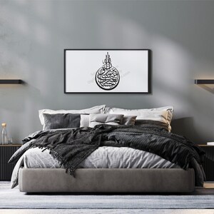 May include: A modern bedroom with a gray wall, a bed with black and white bedding, a black and white framed print with Arabic calligraphy, and two black wall sconces.