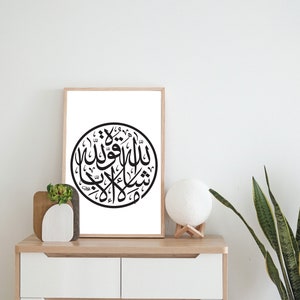 May include: A framed black and white print of Arabic calligraphy in a circle on a white background. The print is hanging on a white wall above a white dresser with two drawers.