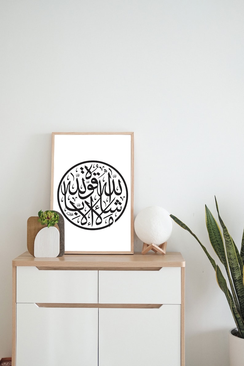 May include: A framed black and white print of Arabic calligraphy in a circle on a white background. The print is hanging on a white wall above a white dresser with two drawers.