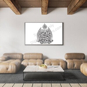 May include: A black and white Islamic calligraphy art print in a black frame hanging on a white wall above a brown leather sectional sofa. The print features a decorative design with Arabic script.