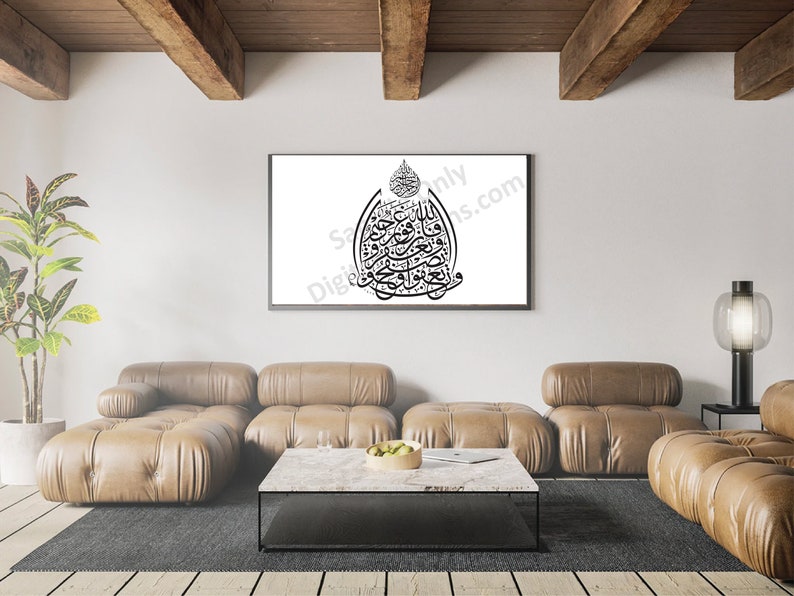 May include: A black and white Islamic calligraphy art print in a black frame hanging on a white wall above a brown leather sectional sofa. The print features a decorative design with Arabic script.
