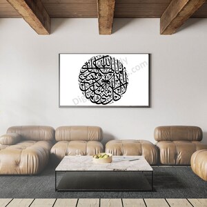 May include: A black and white calligraphy print of Islamic text in a circular frame, hanging on a white wall in a living room setting. The print is framed in a black frame and is hanging above a brown leather sofa and a coffee table.
