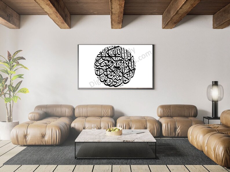 May include: A black and white calligraphy print of Islamic text in a circular frame, hanging on a white wall in a living room setting. The print is framed in a black frame and is hanging above a brown leather sofa and a coffee table.