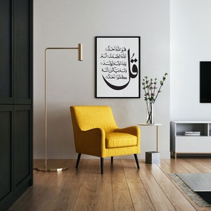 May include: A yellow armchair with black legs sits in a living room with a white wall, a black and white framed print with Arabic calligraphy, a gold floor lamp, a small white table with a vase of pink flowers, and a white entertainment center with a black TV.