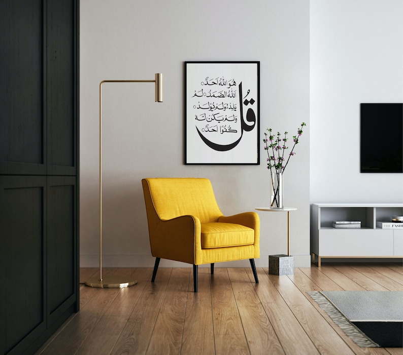May include: A yellow armchair with black legs sits in a living room with a white wall, a black and white framed print with Arabic calligraphy, a gold floor lamp, a small white table with a vase of pink flowers, and a white entertainment center with a black TV.