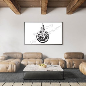 May include: A black and white calligraphy print of Islamic text in a circular frame, hanging on a white wall in a living room setting. The print is framed and is above a brown leather couch and a coffee table.