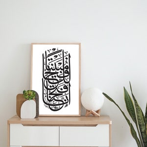 May include: A black and white calligraphy print in a wooden frame, hanging on a white wall above a white dresser with two drawers. A small potted plant and a white lamp are on the dresser.