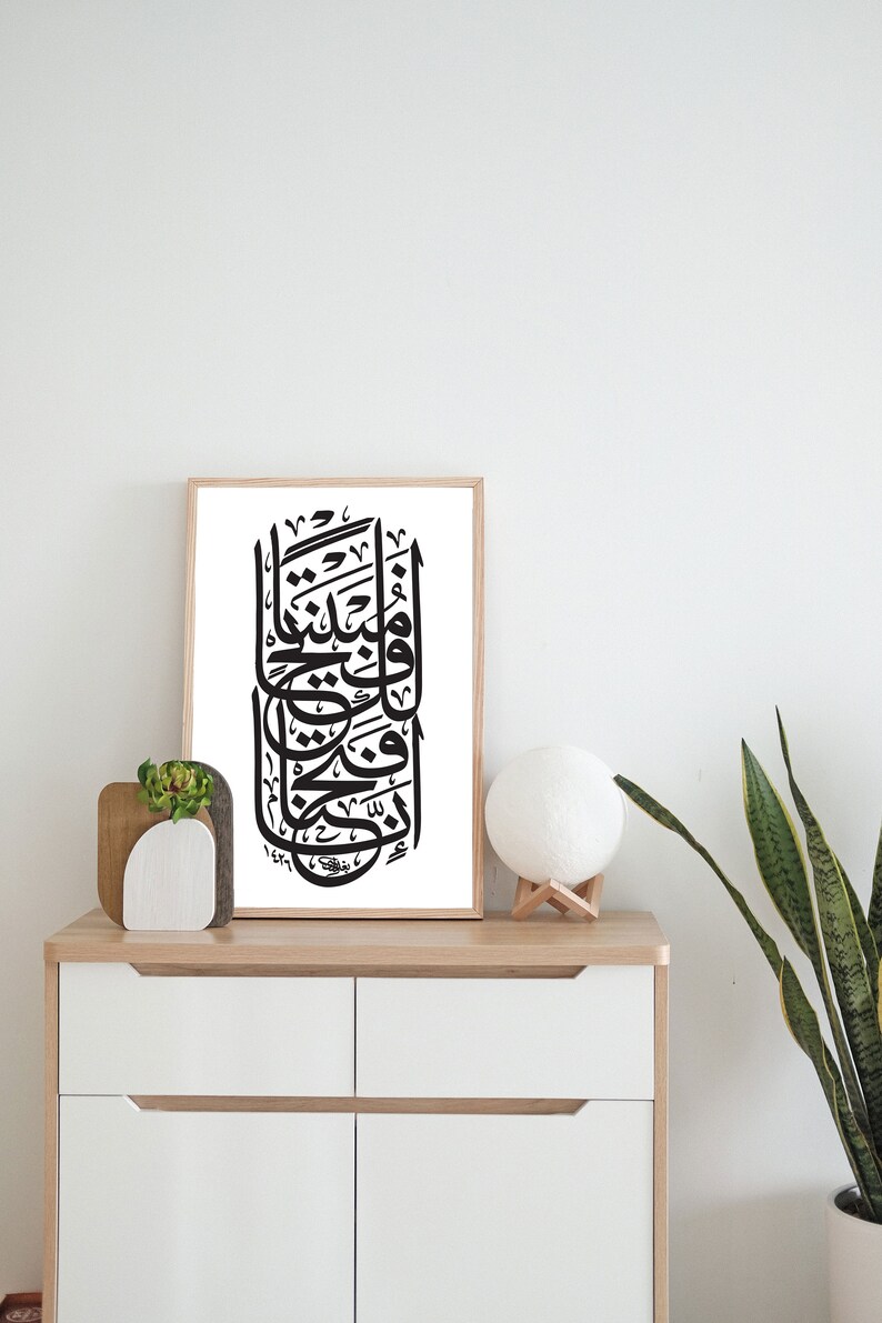 May include: A black and white calligraphy print in a wooden frame, hanging on a white wall above a white dresser with two drawers. A small potted plant and a white lamp are on the dresser.