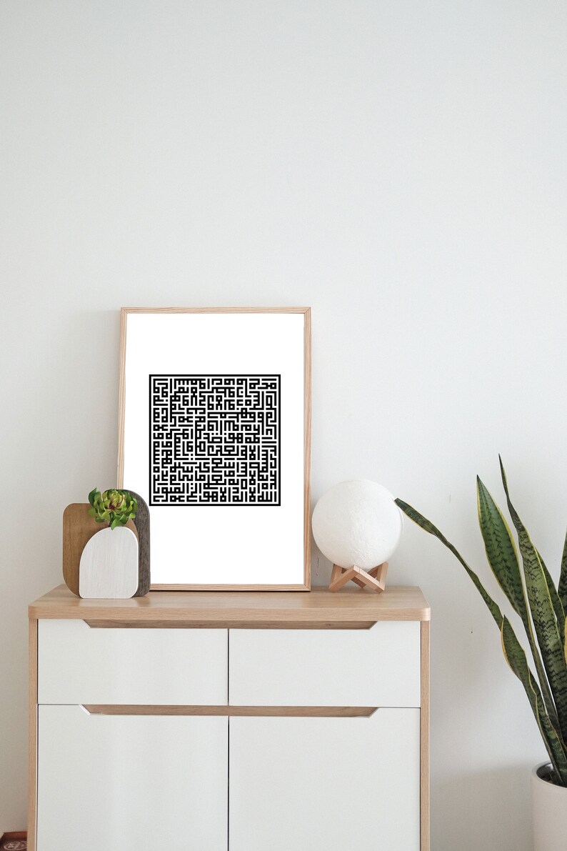 May include: A black and white framed print of Islamic calligraphy in a square shape. The print is hanging on a white wall above a white dresser with two drawers. A small potted plant and a white lamp are on the dresser.