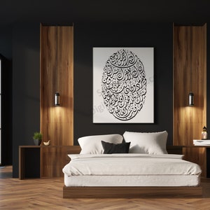 Op de afbeelding: Een wit bed met een wit dekbedovertrek en zwarte kussens staat in een moderne slaapkamer met donkere muren en houten accenten. Een grote zwart-witprint van Arabische kalligrafie hangt aan de muur boven het bed.