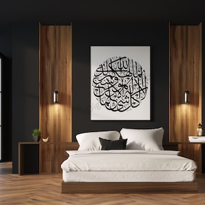 May include: A black and white calligraphy artwork of Islamic text hangs above a white bed with black and white pillows in a modern bedroom with dark walls and wood paneling.