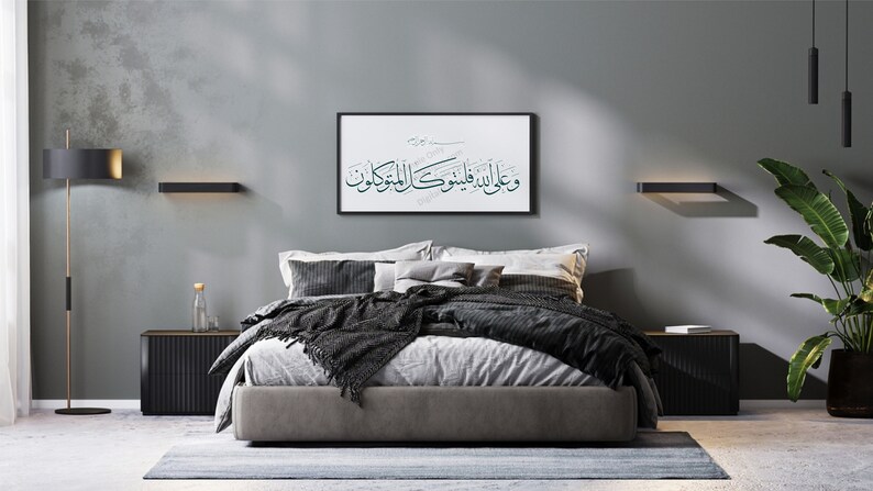 Op de afbeelding: Een slaapkamer met een grijze muur, een bed met een grijs hoofdeinde en zwarte beddengoed, een zwart nachtkastje, een zwarte staande lamp en een ingelijst afdruk met Arabische kalligrafie die "Wa alaa alladhina amanoo wa amiloo al-salihati" luidt
