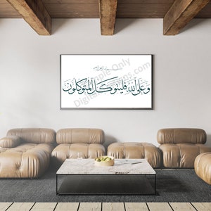 Op de afbeelding: Een ingelijst afdruk van Arabische kalligrafie in zwart en turquoise op een witte achtergrond, hangend aan een witte muur in een woonkamer. De kamer heeft een bruine leren modulaire bank, een salontafel, een kamerplant en een lamp.