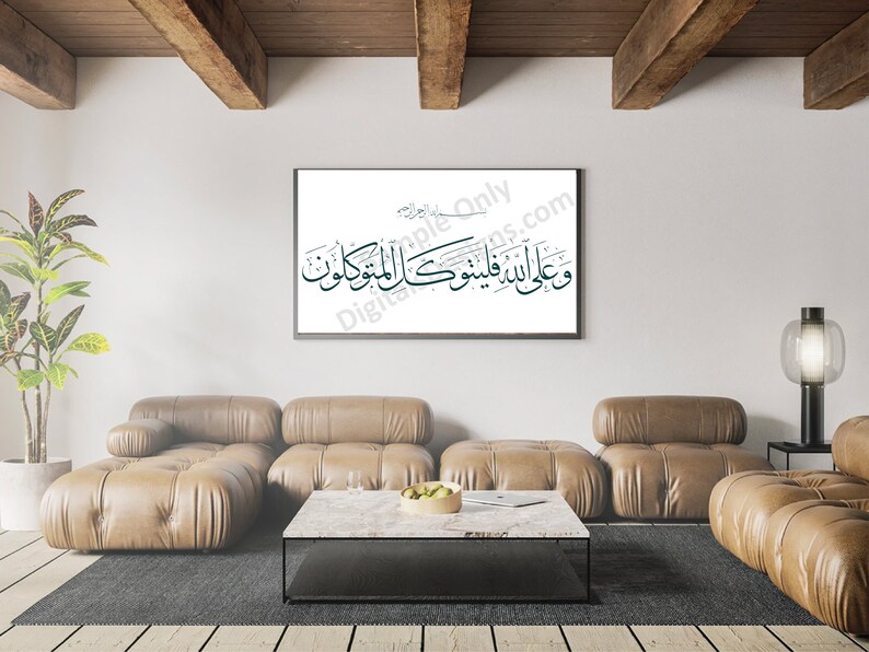 Op de afbeelding: Een ingelijst afdruk van Arabische kalligrafie in zwart en turquoise op een witte achtergrond, hangend aan een witte muur in een woonkamer. De kamer heeft een bruine leren modulaire bank, een salontafel, een kamerplant en een lamp.