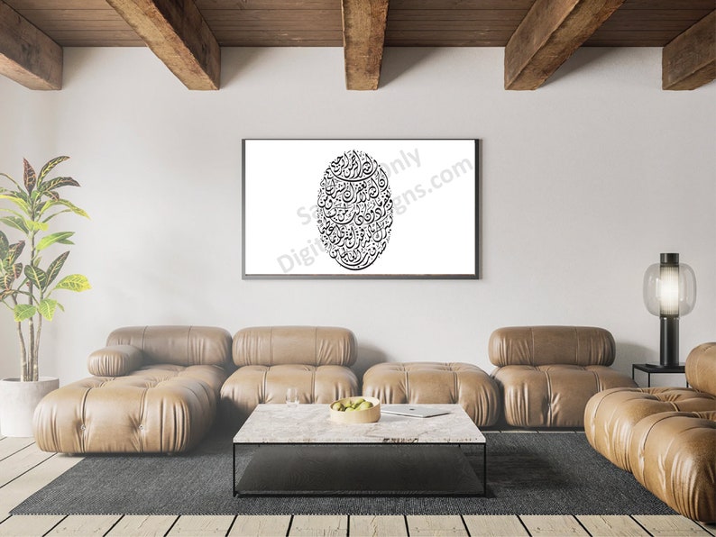 Op de afbeelding: Een zwart-wit kalligrafie kunstprint in een zwart frame hangend aan een witte muur boven een bruine leren sectionele bank. De bank staat in een woonkamer met een houten vloer en een salontafel met een fruitschaal erop.