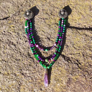 May include: A multi-strand necklace featuring green, purple, and pink beads, with silver-toned decorative clasps. A clear, elongated crystal pendant hangs from the bottom. The necklace is displayed on a textured, light brown surface.