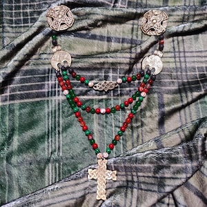 以下が含まれることがあります： 金色のケルト十字ペンダントと装飾的な金属製の留め金が付いた、マルチストランドネックレス。 ネックレスは赤、緑、白のビーズを組み込み、模様のある背景があります。