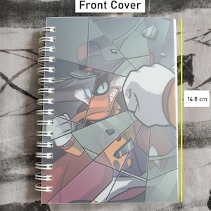 Puede incluir: Un cuaderno de espiral con una ilustración colorida de un personaje de dibujos animados rompiendo una ventana. La portada mide 10,5 cm de ancho y 14,8 cm de alto.