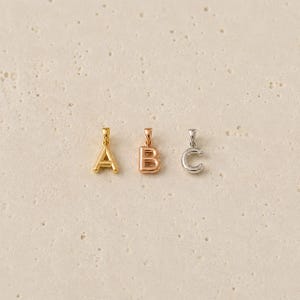 May include: Three individual letter pendants arranged in a row. The first is a gold-toned letter A, the second is a rose gold-toned letter B, and the third is a silver-toned letter C. Each pendant features a small integrated bail at the top.