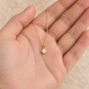 May include: A gold chain necklace featuring a small, round, iridescent white opal pendant set in a gold bezel with a delicate beaded edge. The pendant hangs from a thin, polished gold cable chain, creating a simple and refined jewelry piece.