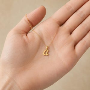 May include: A gold-toned necklace featuring a small, lowercase letter 'a' pendant suspended from a delicate, thin chain. The pendant has a smooth, polished finish and is centered on the chain, which rests against the palm of an open hand.