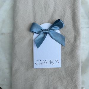 May include: A white tag with the name "CAMERON" in elegant script is decorated with a blue satin bow. The tag is placed on a beige linen napkin with a fringed edge, creating a simple yet refined aesthetic.