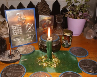 May include: A green table with a lit candle and several silver and copper plates with blue trim. The plates have symbols and Hebrew writing on them. There is a small glass jar with a cork lid and a metal plate with numbers and symbols on it. The table is covered with a green cloth.