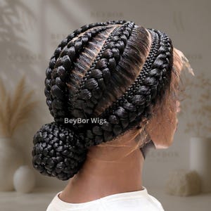 May include: A black wig featuring intricate cornrow braids that sweep back from the hairline and gather into a low, rounded bun at the nape of the neck. The lace front is visible along the hairline, and the text BeyBor Wigs is printed on the side.