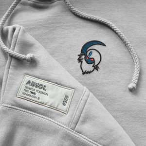 May include: A pale grey hoodie featuring a drawstring and a small embroidered design of a creature with blue, white, and red accents. The hoodie has a rectangular patch with the text "Absol Disaster Pokemon Type Dark Generation III" and the number "#359".