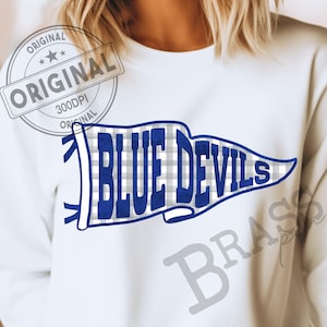 May include: A white crewneck sweatshirt featuring a graphic of a pennant flag with a gray and white gingham pattern. The text "BLUE DEVILS" is printed in bold blue letters across the center of the pennant. The design includes blue outlines.