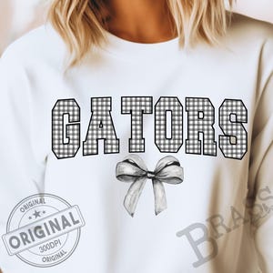 May include: White sweatshirt with the word "GATORS" in large, black and white checkered letters. A gray bow is below the word. The word "ORIGINAL" is printed on a gray stamp.
