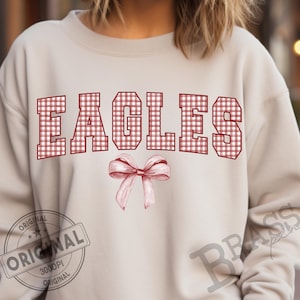 May include: A light beige sweatshirt with the word "EAGLES" in red and white gingham block letters. A watercolor-style pink bow is centered below the word. The word "ORIGINAL" is printed on a stamp.