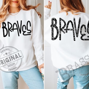May include: White long-sleeved sweatshirt with the word "BRAVES" printed in large, black, stylized letters on the front and back. The sweatshirt has a crew neck and is paired with blue jeans. A circular stamp with the word "ORIGINAL" is visible.