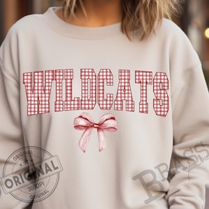 May include: A light beige sweatshirt with the word "WILDCATS" in red and white gingham print across the chest. A watercolor-style bow is below the text. The sweatshirt has a crew neck and long sleeves.