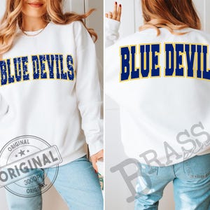 Puede incluir: Sudadera blanca con el texto "BLUE DEVILS" en azul marino con un contorno amarillo en la parte delantera y trasera. La sudadera se combina con unos vaqueros azules.