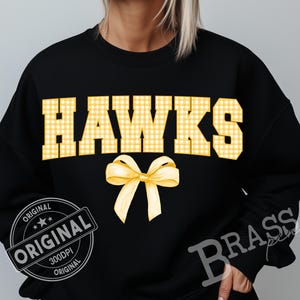 May include: Black sweatshirt with the word "HAWKS" in yellow gingham block letters and a yellow bow graphic. The sweatshirt also has a "ORIGINAL" stamp and "Brass" text.