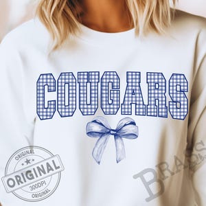 Puede incluir: Sudadera blanca con la palabra "COUGARS" en estampado de cuadros vichy azules. Un lazo azul está debajo de la palabra. La palabra "ORIGINAL" está impresa en un sello circular.