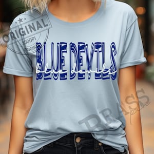 May include: A light blue t-shirt with the words "BLUE DEVILS" in a bold, navy blue font. The words "blue devils" are also written in a cursive white font. The shirt is a short-sleeved crew neck.