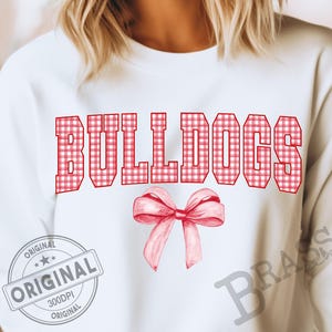 Puede incluir: Sudadera blanca con la palabra "BULLDOGS" en letras rojas a cuadros. Debajo del texto hay un lazo rojo estilo acuarela. La sudadera tiene cuello redondo y mangas largas.