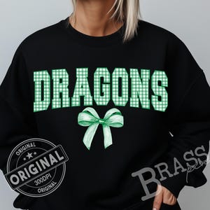 May include: Black sweatshirt with the word "DRAGONS" in large, green gingham letters. A green bow is below the word. The sweatshirt has a "ORIGINAL" stamp and the word "BRASS" is printed on the sleeve.