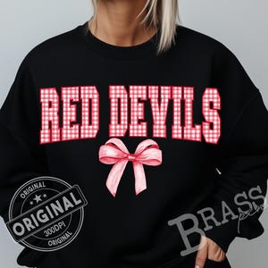 May include: Black sweatshirt with the words "RED DEVILS" in red and white gingham print. A pink bow is below the text. The word "ORIGINAL" is printed on a black and white stamp.