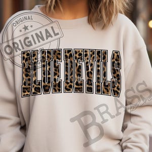 May include: A beige sweatshirt with the words "BLUE DEVILS" in large, bold letters across the front. The letters have a leopard print pattern. The word "ORIGINAL" is stamped on the upper left side.