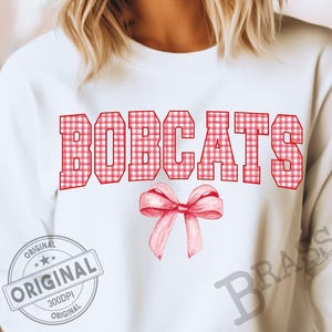 Puede incluir: Sudadera blanca con la palabra "BOBCATS" en letras de cuadros rojos. Una cinta rosa está debajo del texto. La sudadera tiene un aspecto vintage con un sello "ORIGINAL".