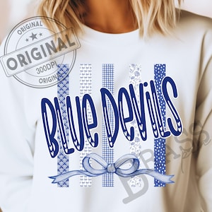 May include: A white sweatshirt featuring a graphic design with the text BLUE DEVILS in dark blue letters. The design includes five vertical stripes with various blue and white patterns, including gingham, polka dots, and floral, with a light blue bow.