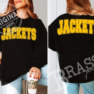 May include: A black long-sleeve sweatshirt featuring the word JACKETS printed in a distressed yellow varsity-style font on the front and a clean yellow varsity-style font on the back. The garment is paired with light blue denim jeans.